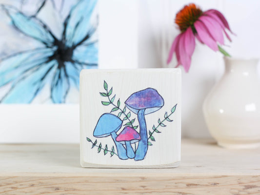 Whitewashed wood block sign with  some whimsical blue, purple, and pink mushrooms on it, intertwined with some vines with green leaves.