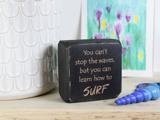 Small distressed black wood sign with the saying "You can't stop the waves, but you can learn to surf"