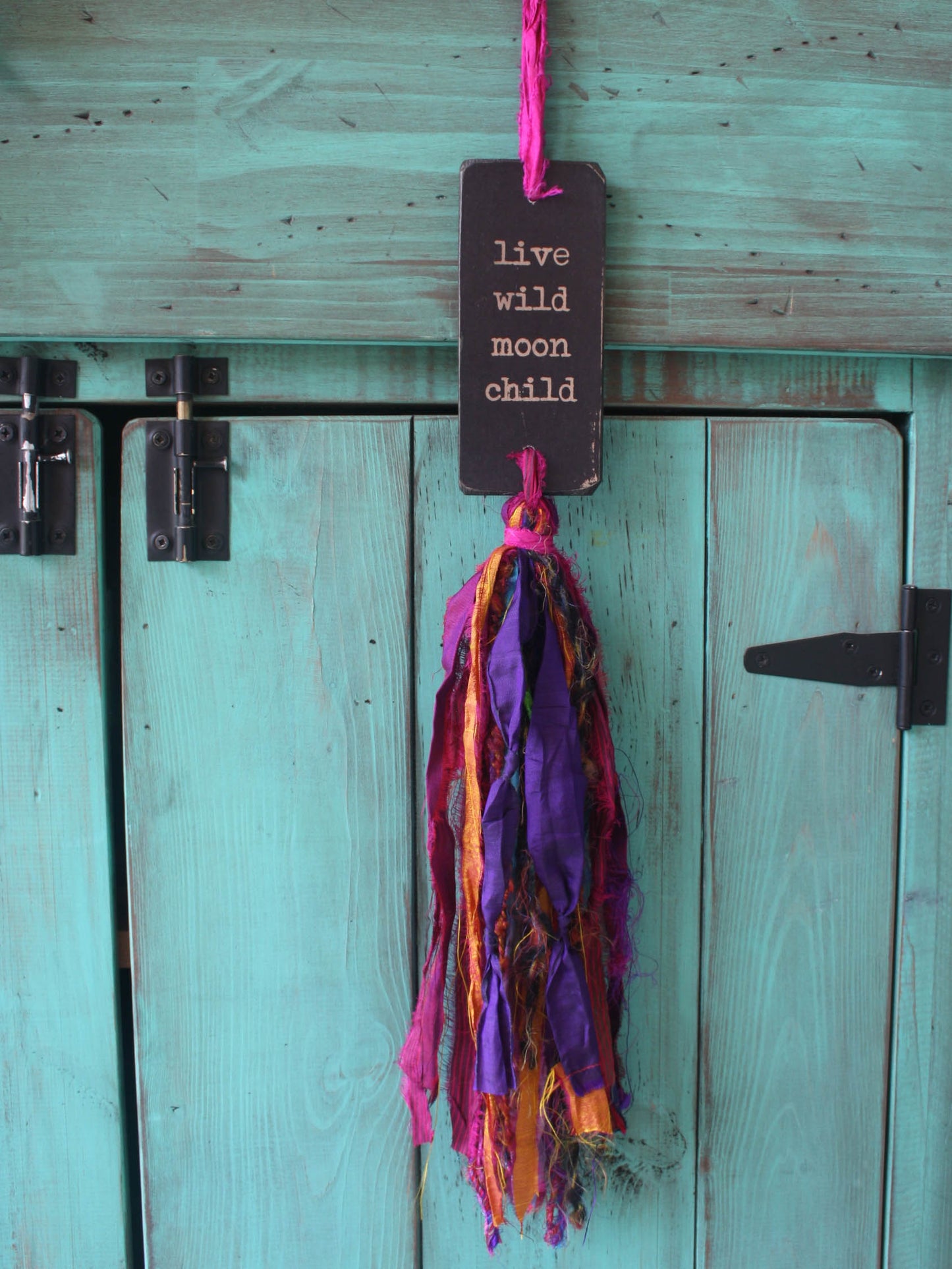 a rectangular piece of wood in distressed black with the text "live wild moon child" and a colourful multicoloured tassel.