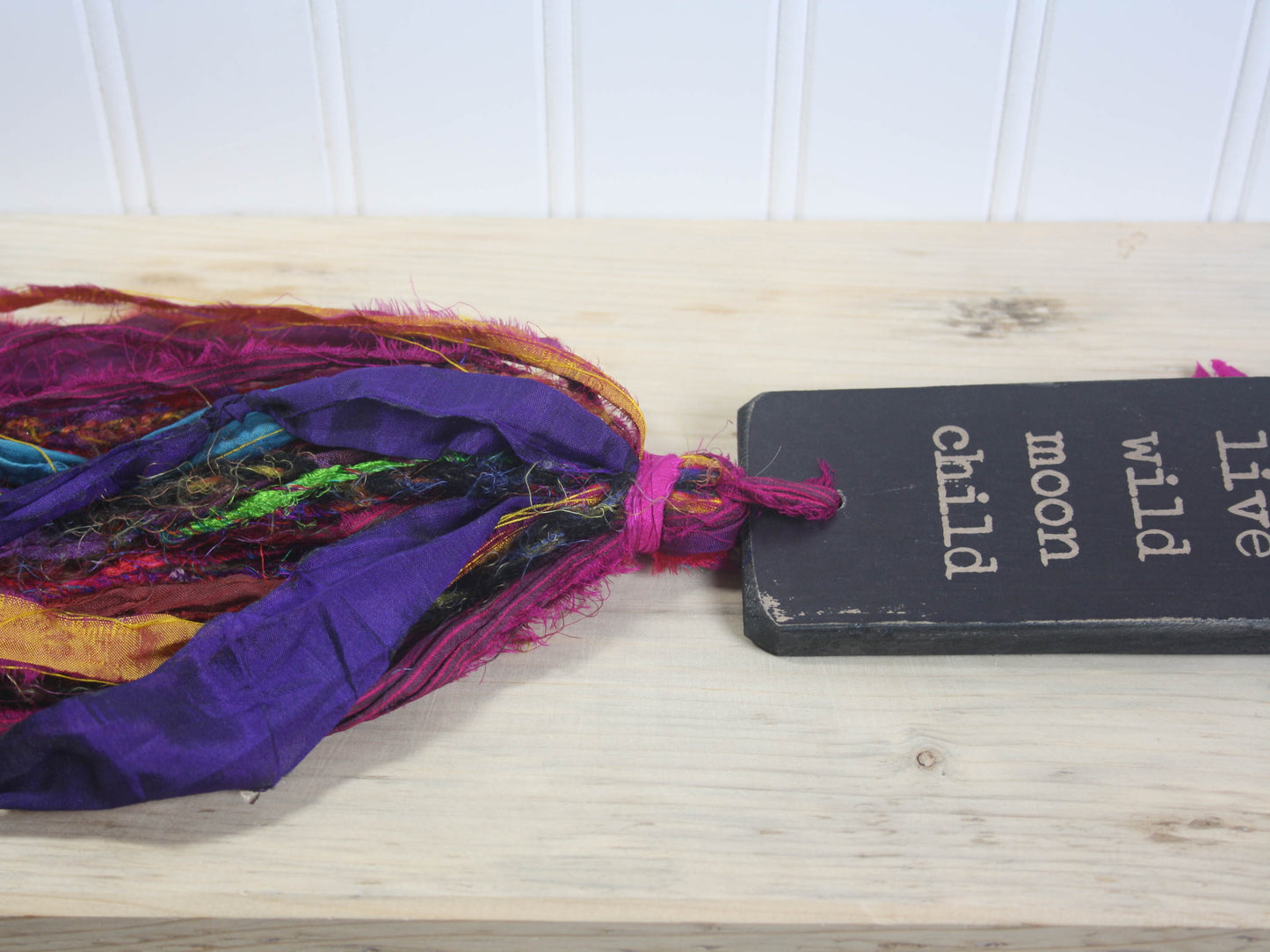 a rectangular piece of wood in distressed black with the text "live wild moon child" and a colourful multicoloured tassel.