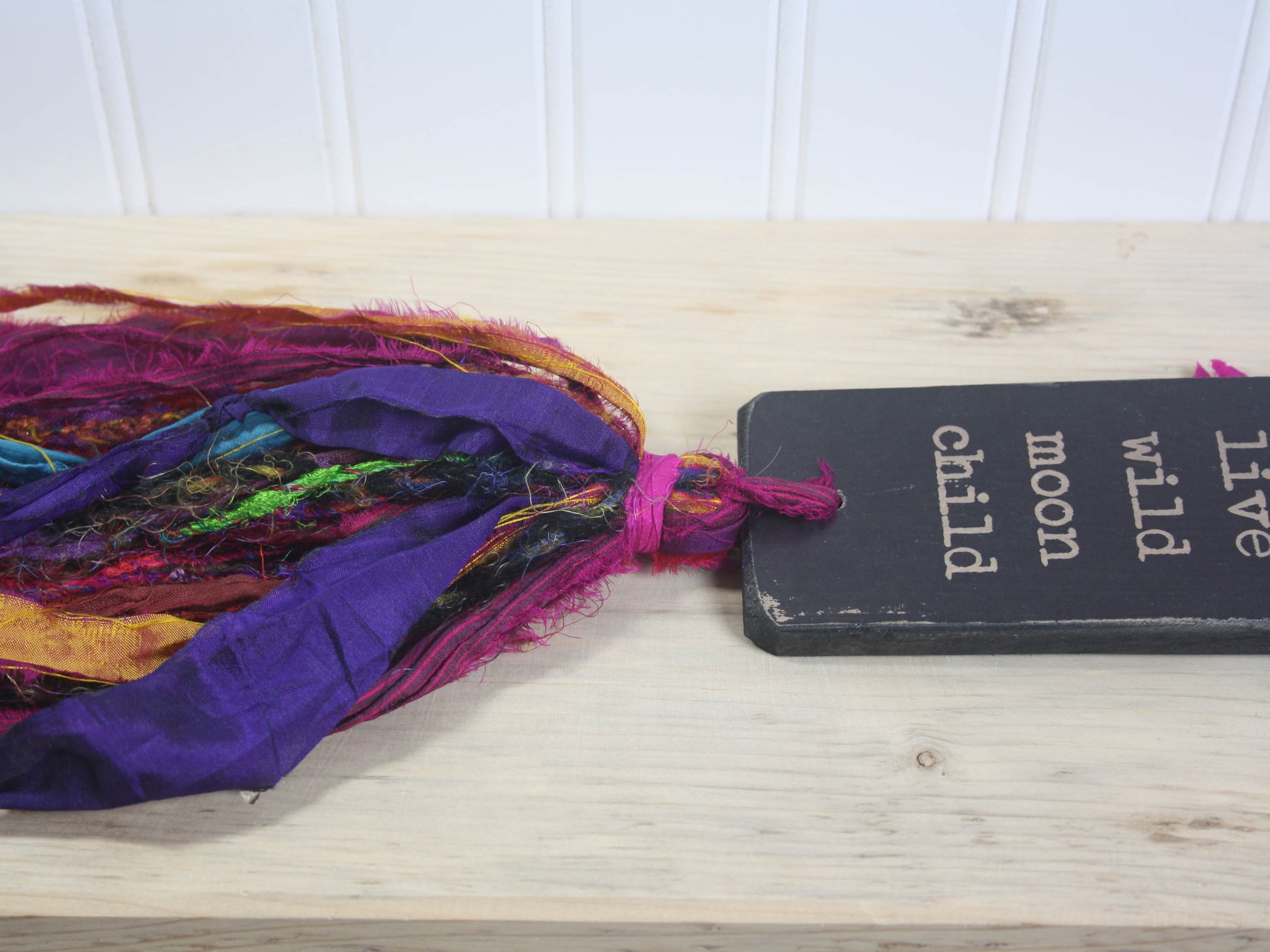 a rectangular piece of wood in distressed black with the text "live wild moon child" and a colourful multicoloured tassel.