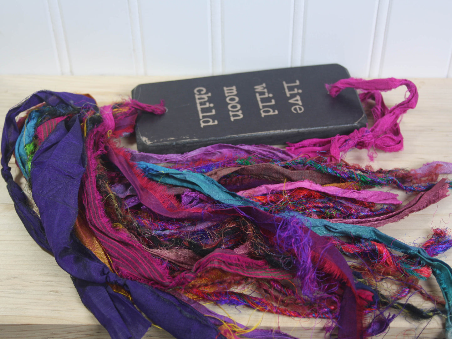 a rectangular piece of wood in distressed black with the text "live wild moon child" and a colourful multicoloured tassel.