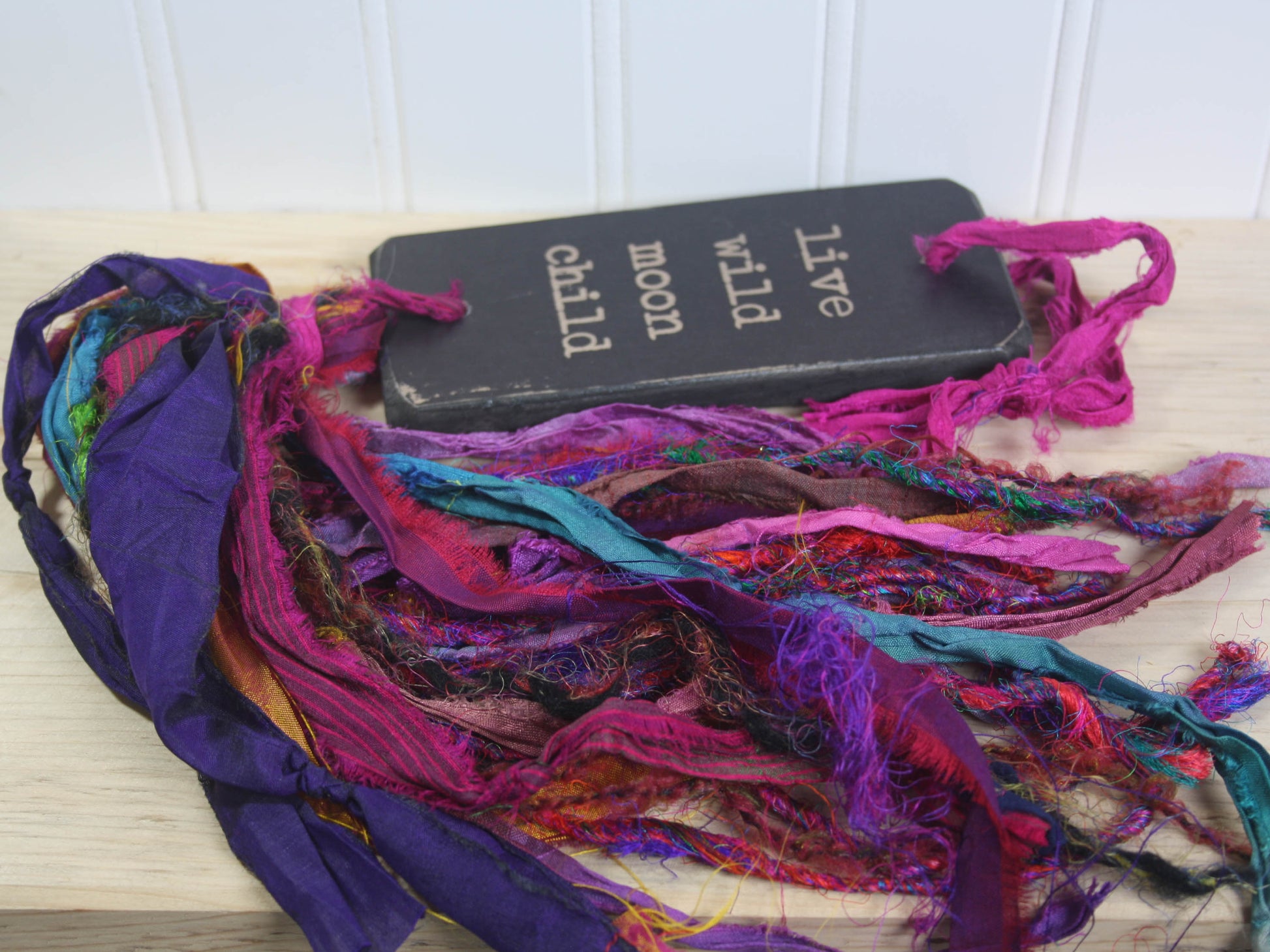 a rectangular piece of wood in distressed black with the text "live wild moon child" and a colourful multicoloured tassel.