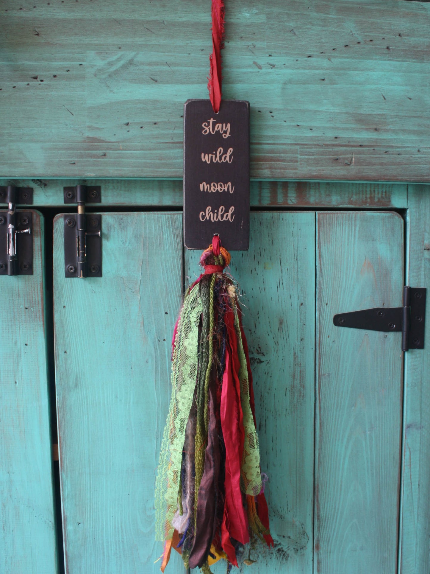 a rectangular piece of wood in distressed black with the text "stay
wild moon child". A colourful multicoloured tassel hangs down from the piece of wood.