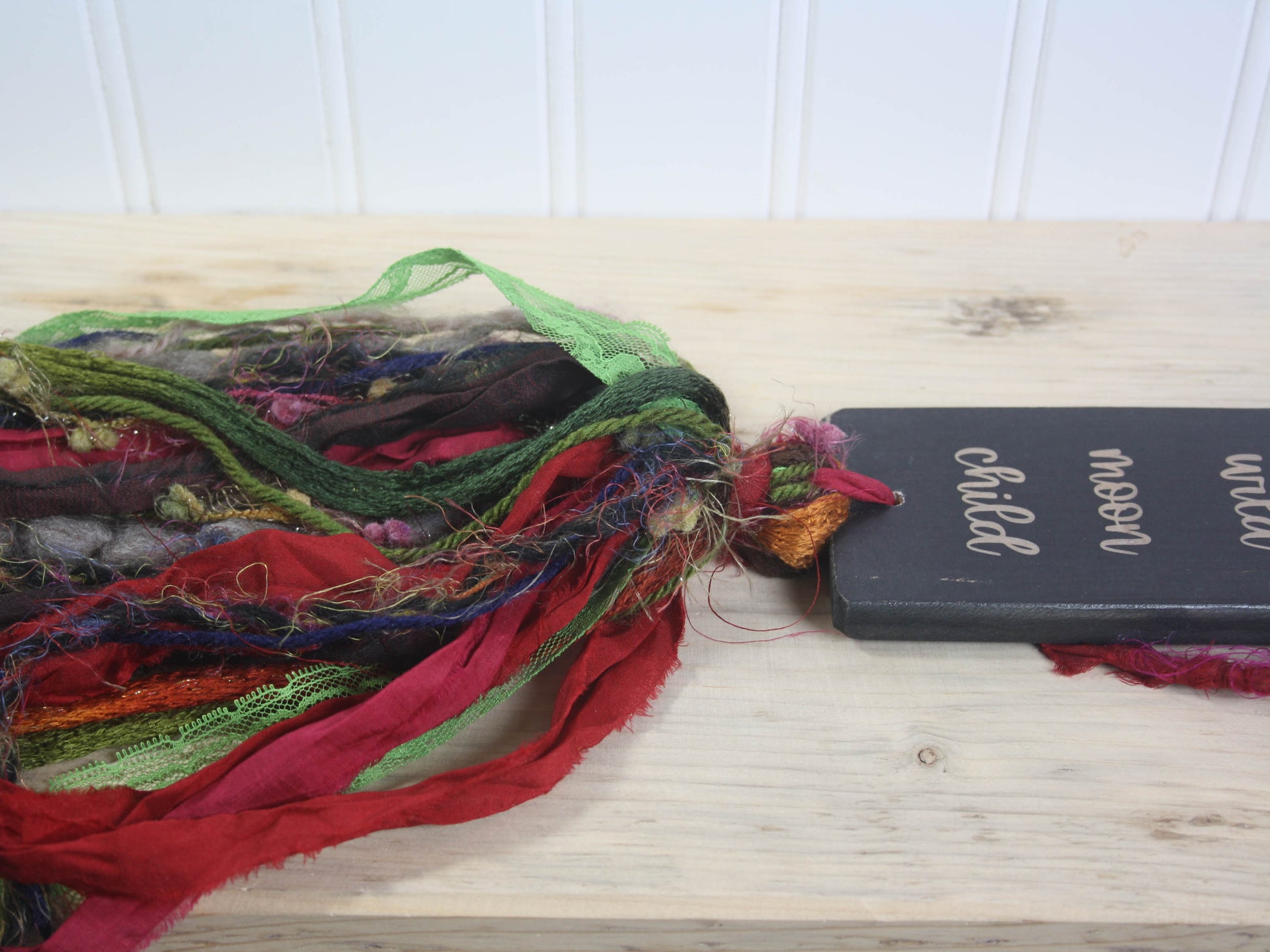 a rectangular piece of wood in distressed black with the text "stay 
 wild moon child". A colourful multicoloured tassel hangs down from the piece of wood.