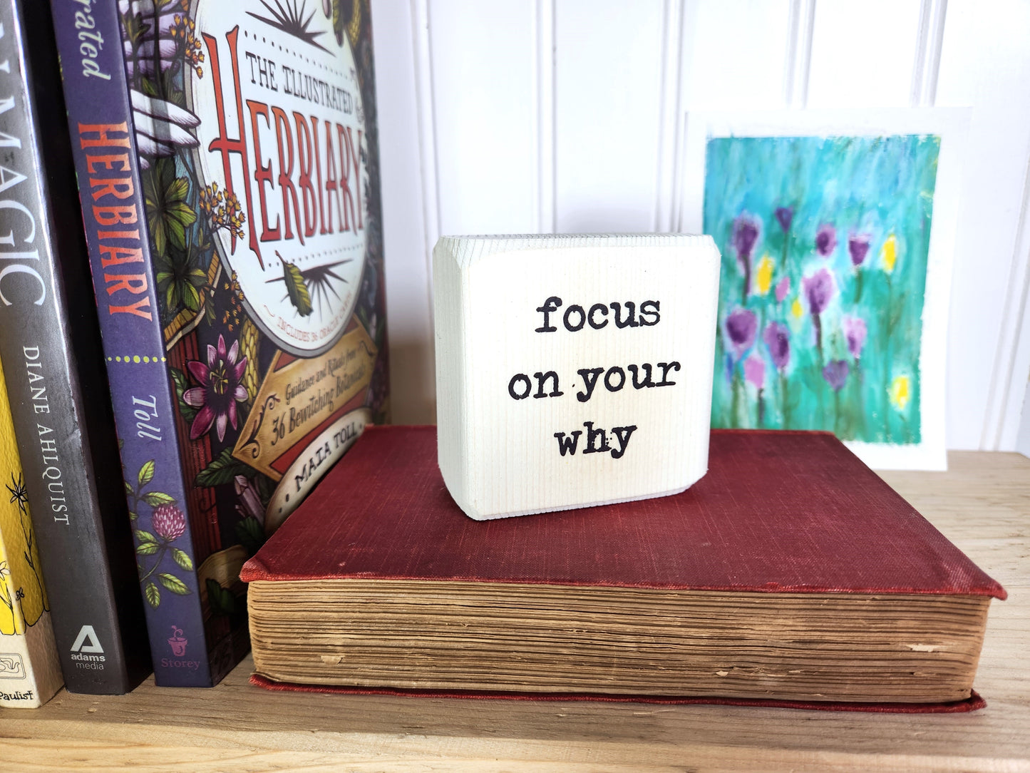 Book with a motivational message on a shelf with other books and a painting.