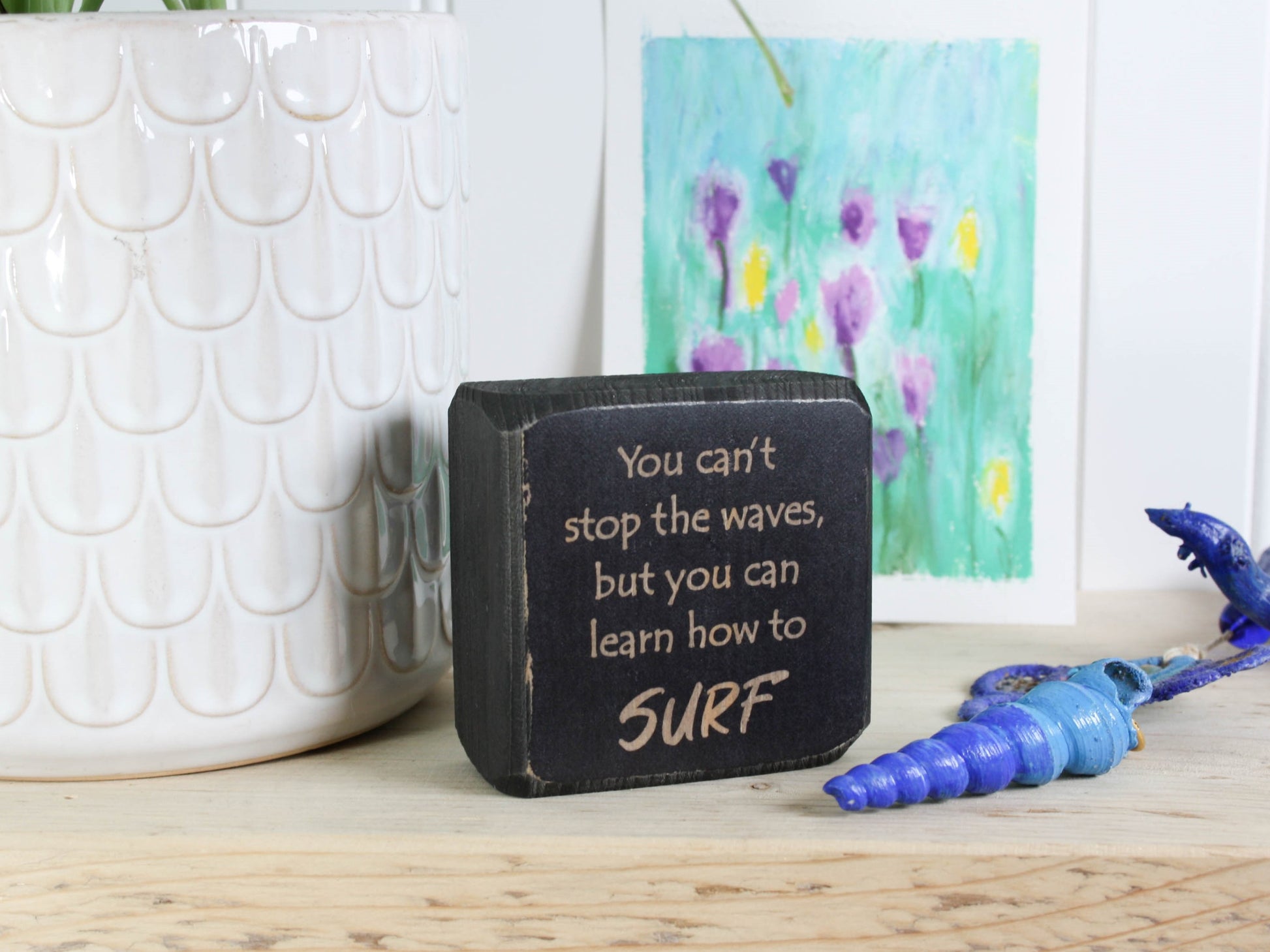 Small distressed black wood sign with the saying "You can't stop the waves, but you can learn to surf"