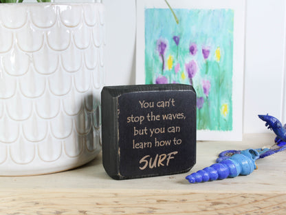 Small distressed black wood sign with the saying "You can't stop the waves, but you can learn to surf"