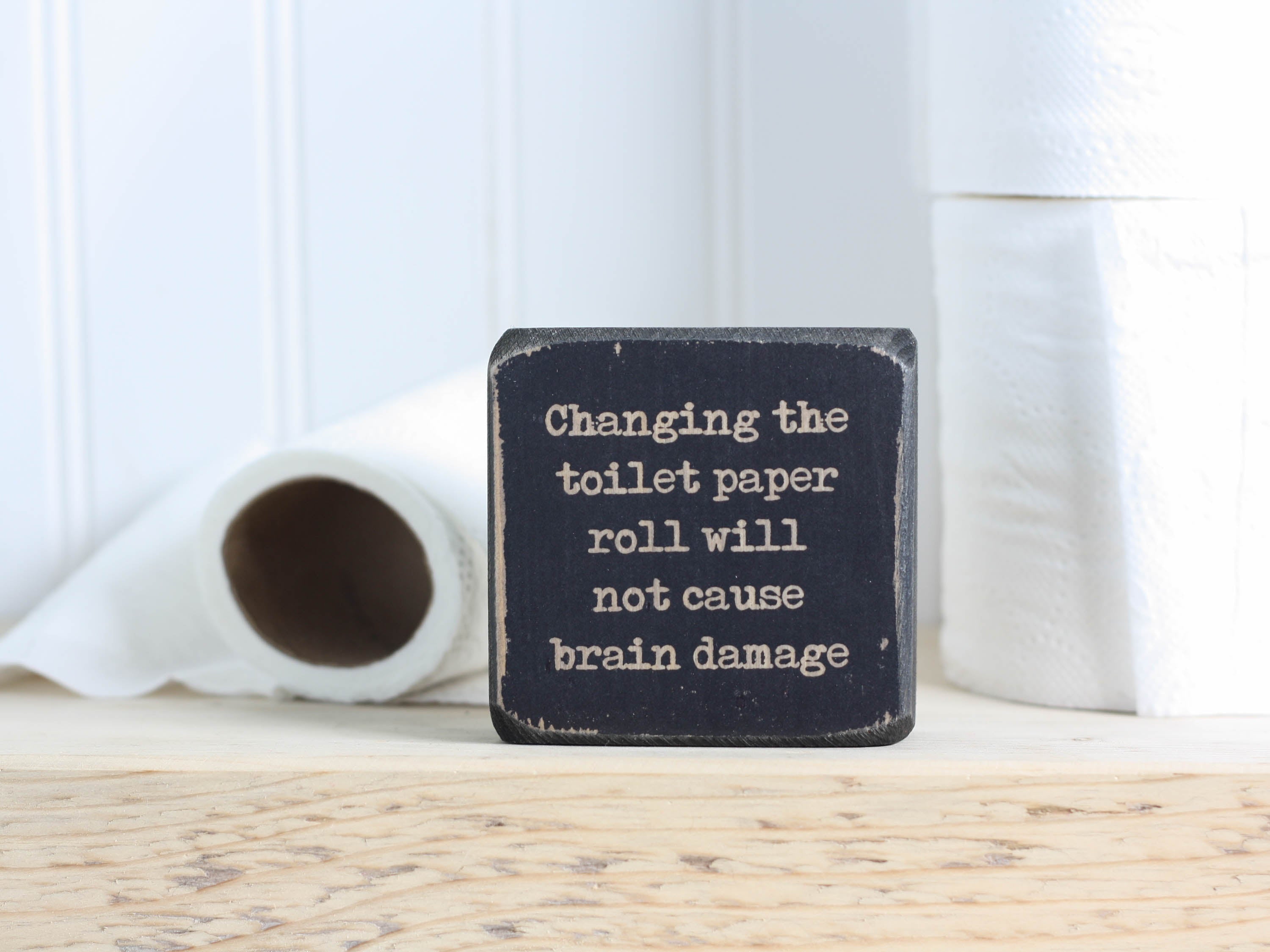 Small wood sign in distressed black with the saying "Changing the toilet paper roll will not cause brain damage."