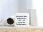 Small wood sign in whitewash with the saying "Changing the toilet paper roll will not cause brain damage."