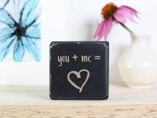 Mini wood desk sign in distressed black with the text "you + me = (heart image)"