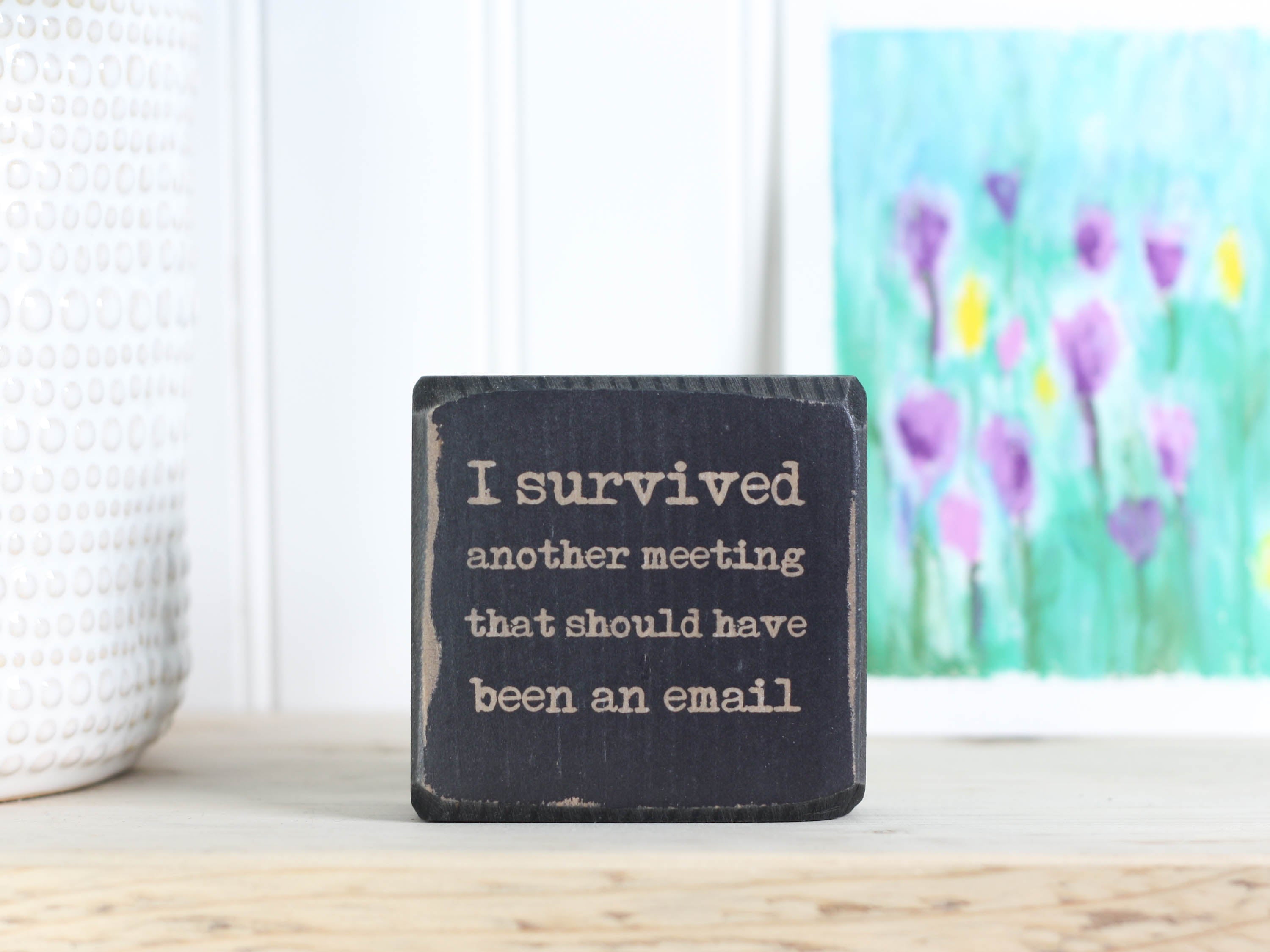 Small, freestanding distressed black wood sign with a funny saying on it "I survived another meeting that should have been an email".