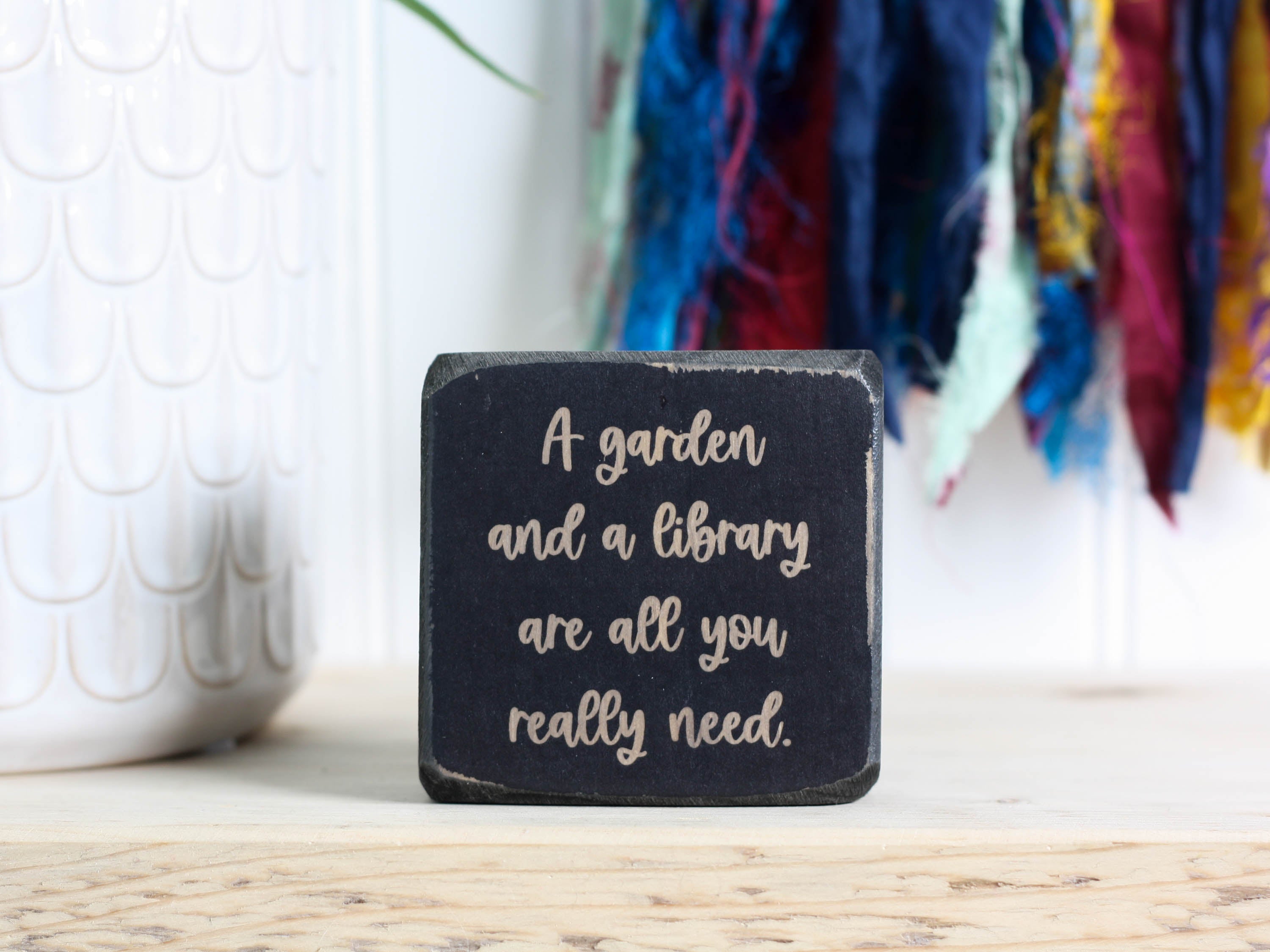 Small wood sign in distressed black with the saying "A garden and a library are all you really need."