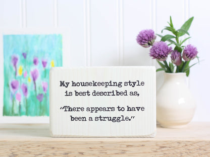 Small funny wood sign in whitewash with the saying "My housekeeping style is best desribed as, "There appears to have been a struggle."