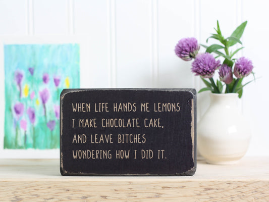 Small free-standing wood sign in distressed black with the saying "When life hands me lemons I make chocolate cake, and leave bitches wondering how I did it.