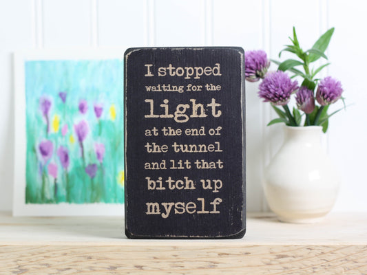 Small wood quote block in distressed black with the saying "I stopped waiting for the light at the end of the tunnel and lit that bitch up myself."