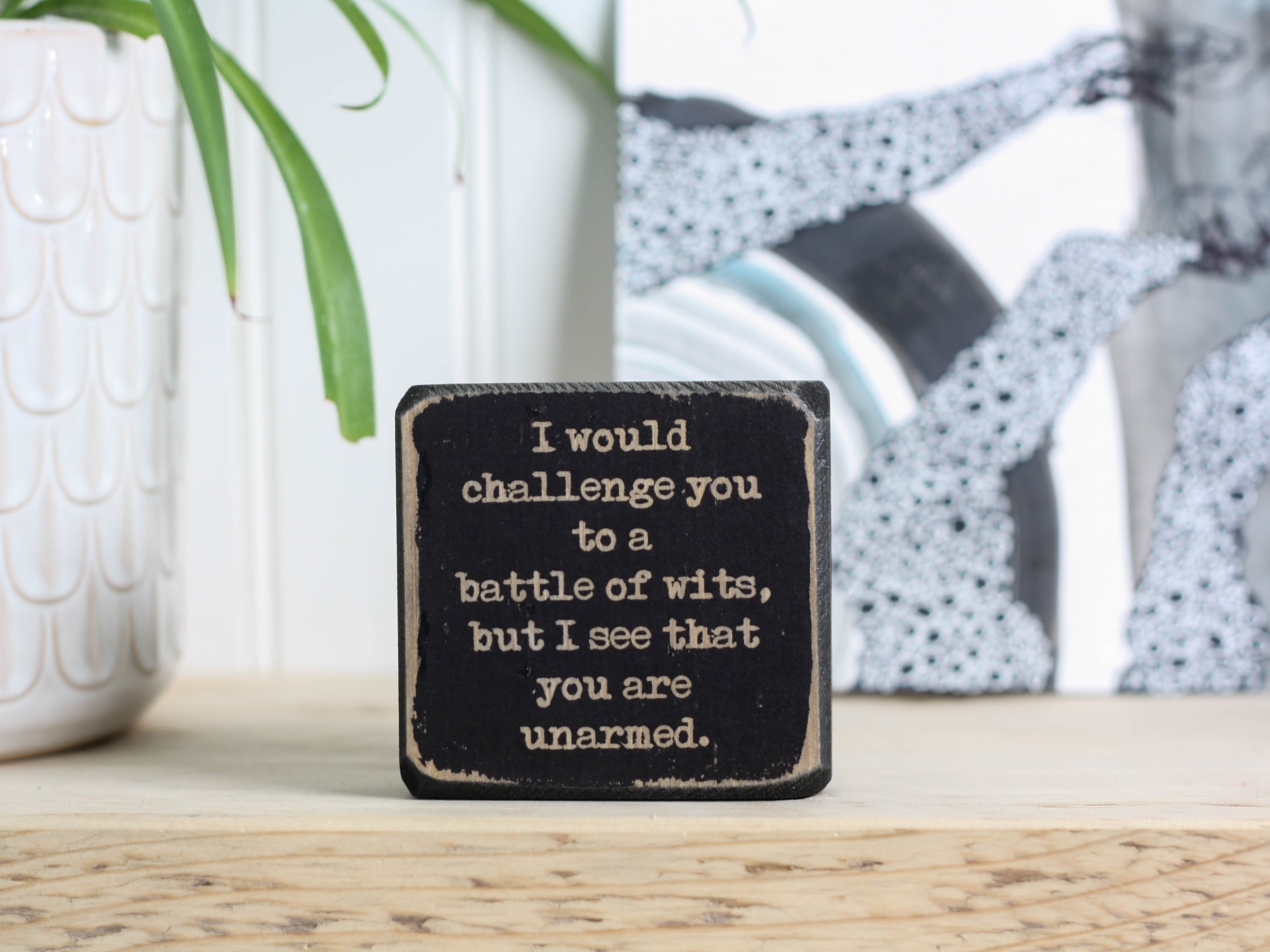 Small wood shelf decor in distressed black with the saying "I would challenge you to a battle of wits, but I see that you are unarmed."