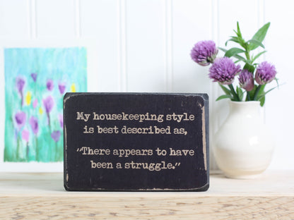 Small funny wood sign in distressed black with the saying "My housekeeping style is best desribed as, "There appears to have been a struggle."