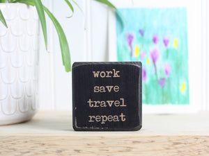 Small wood travel sign in distressed black with the saying "work save travel repeat"
