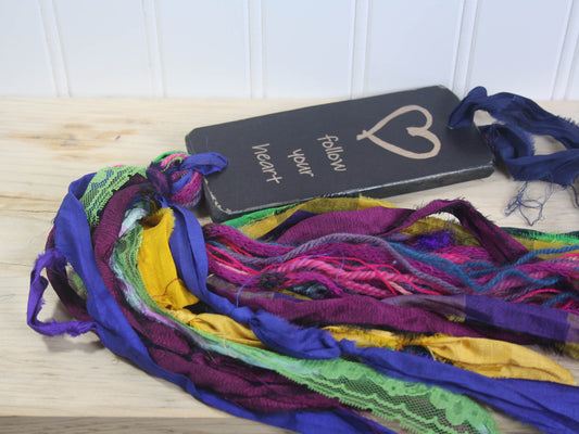 a rectangular piece of wood in distressed black with the text "follow your heart" and an image of a heart. a colourful multicoloured tassel hangs down from the piece of wood.