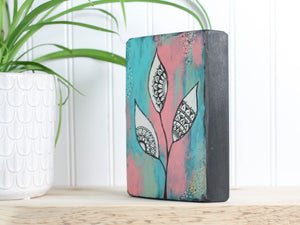 Original mixed media art on a wood block, with a background of teal, peach and dusty light green, with white leaves and mandala inspired shapes inside the leaves.