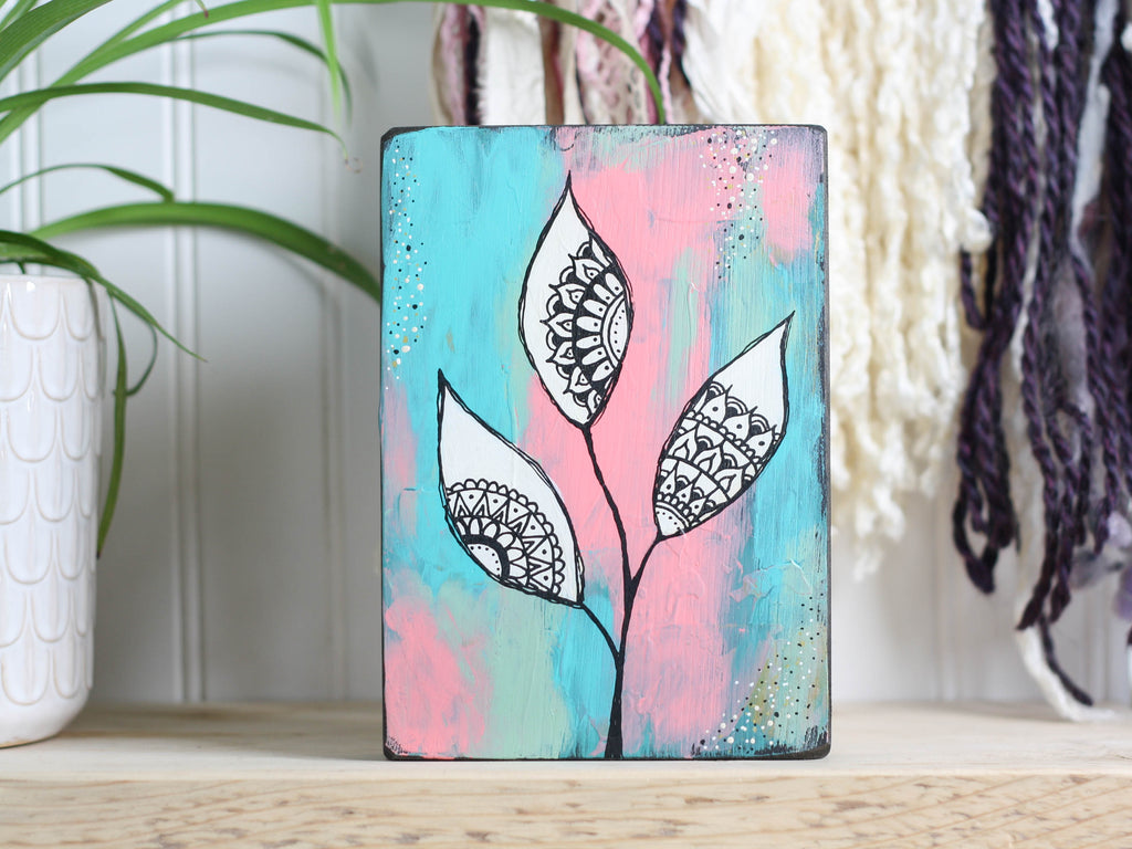 Original mixed media art on a wood block,  with a background of teal, peach and dusty light green, with white leaves and mandala inspired shapes inside the leaves. 