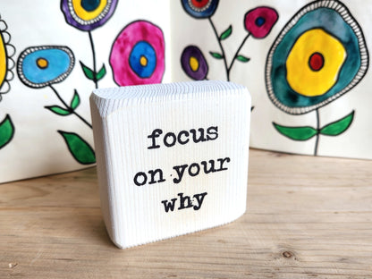 White block sign with 'focus on your why' text in front of colorful floral artwork