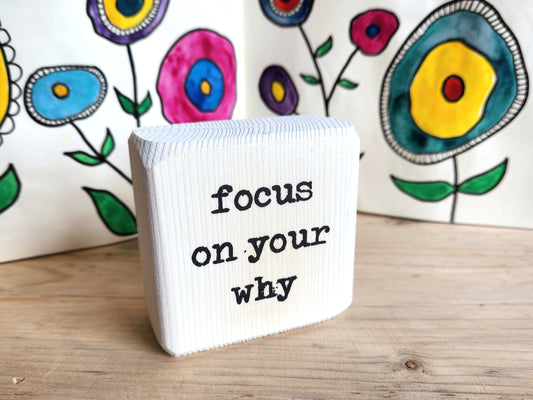 White block sign with 'focus on your why' text in front of colorful floral artwork
