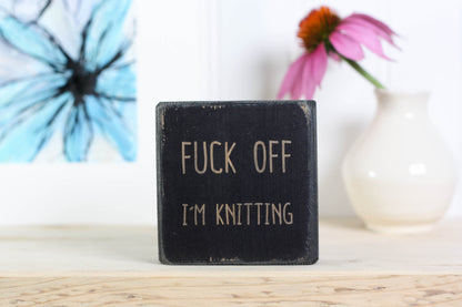 Small distressed black wood sign with the saying "fuck off I'm knitting"