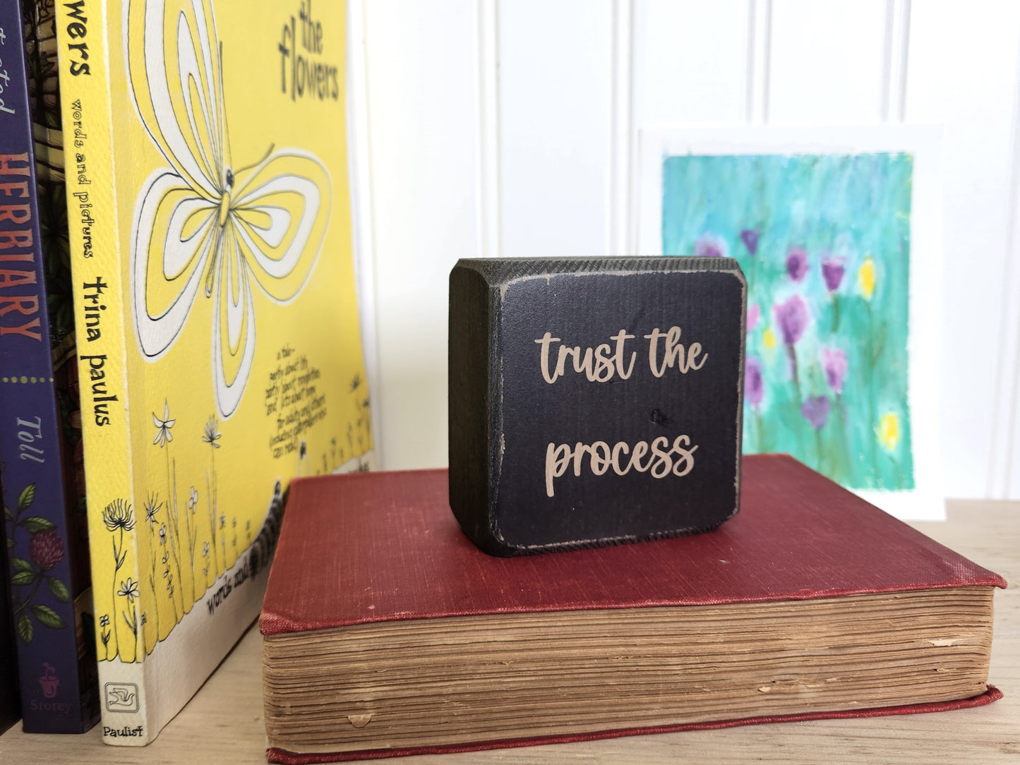 Small black block sign with 'Trust the process' text on a red book, with books and a painting in the background.
