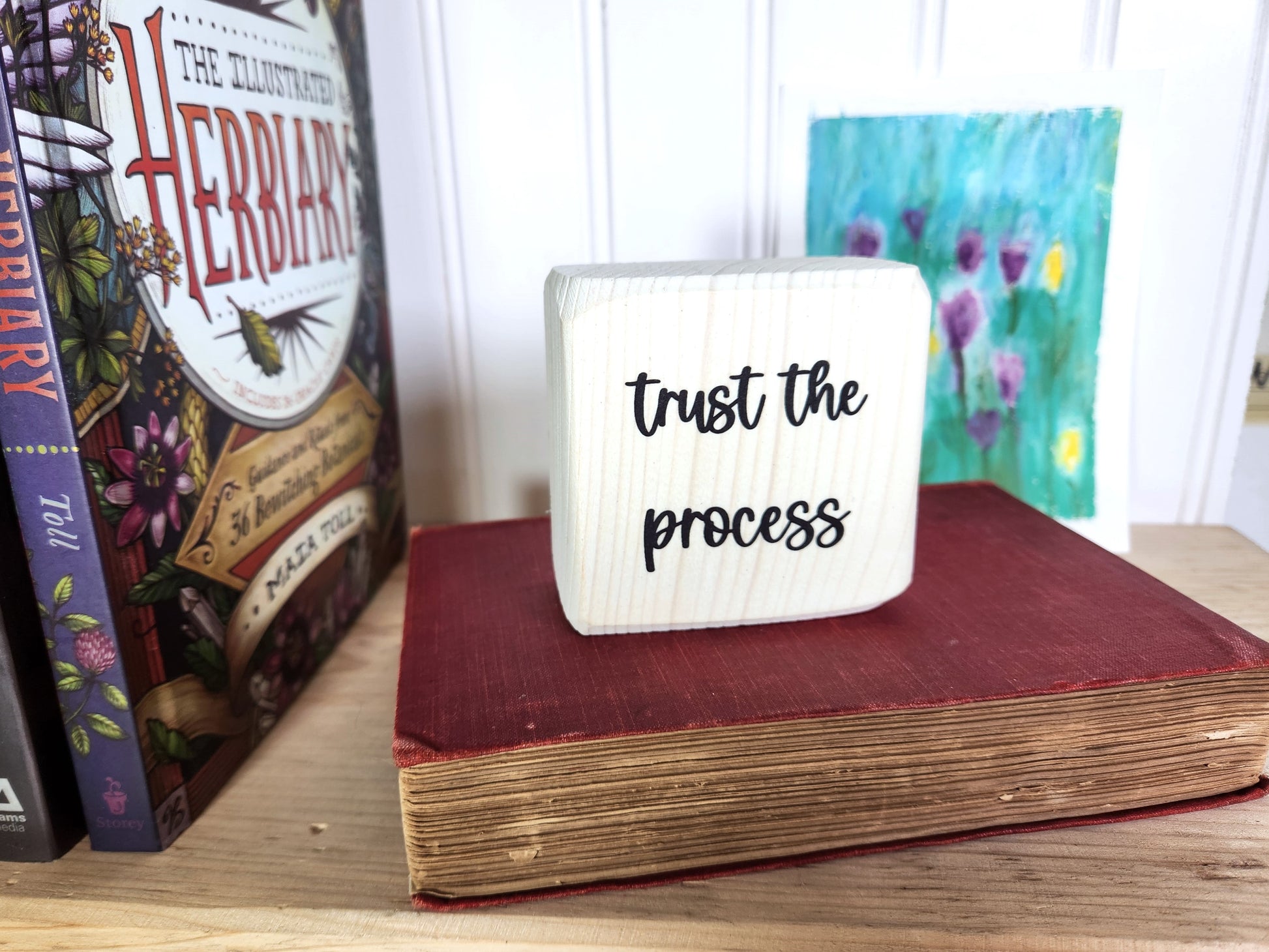 Small white block sign with 'trust the process' text on a wooden bookshelf with books and a painting in the background.