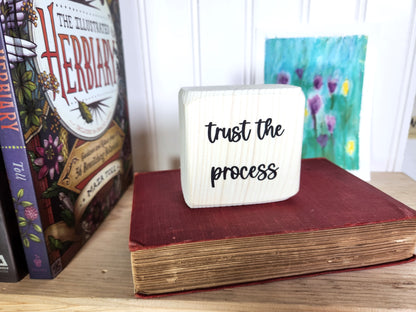 Small white block sign with 'trust the process' text on a wooden bookshelf with books and a painting in the background.