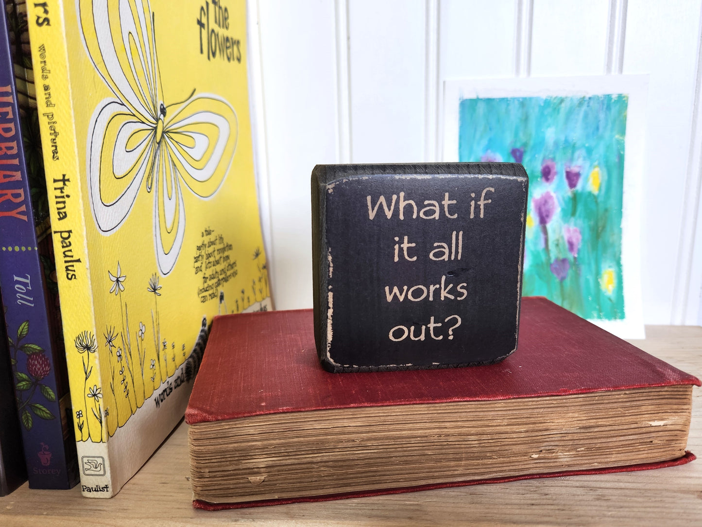 Small black block sign with 'What if it all works out?' text on a red book with books and a painting in the background.