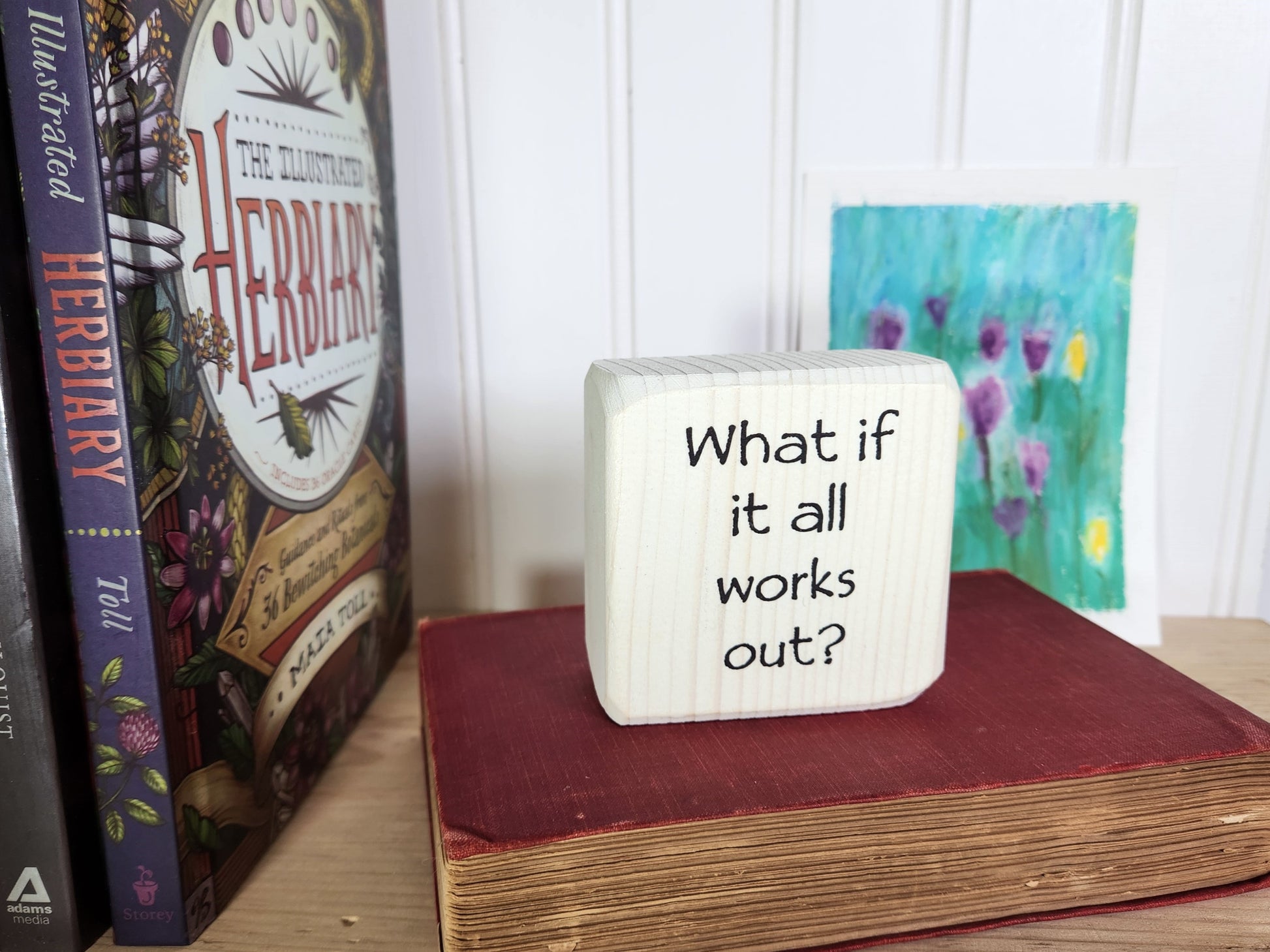 Small whitewashed block sign with 'What if it all works out?' text on a bookshelf with books and a painting in the background.