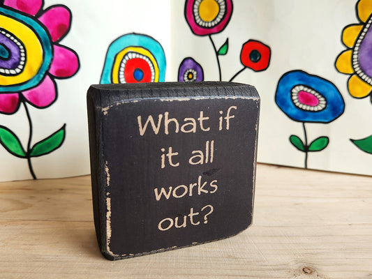 Black sign with 'What if it all works out?' text in front of colorful flower drawings.