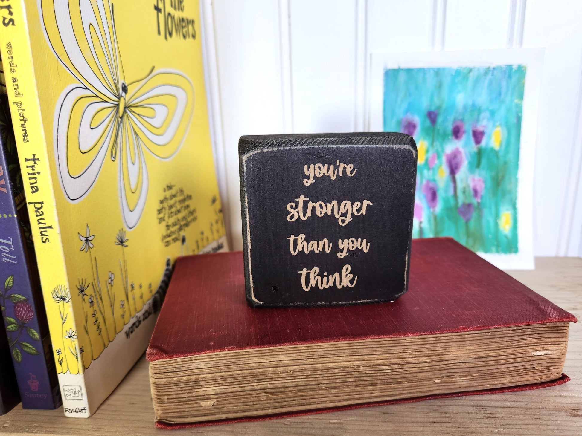 Small black block sign with 'you're stronger than you think' text on a red book with books and a painting in the background.