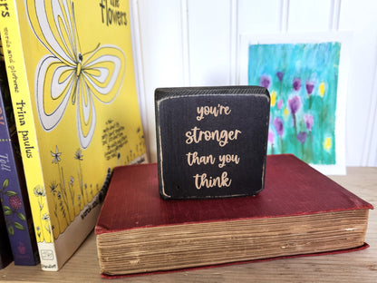 Small black block sign with 'you're stronger than you think' text on a red book with books and a painting in the background.