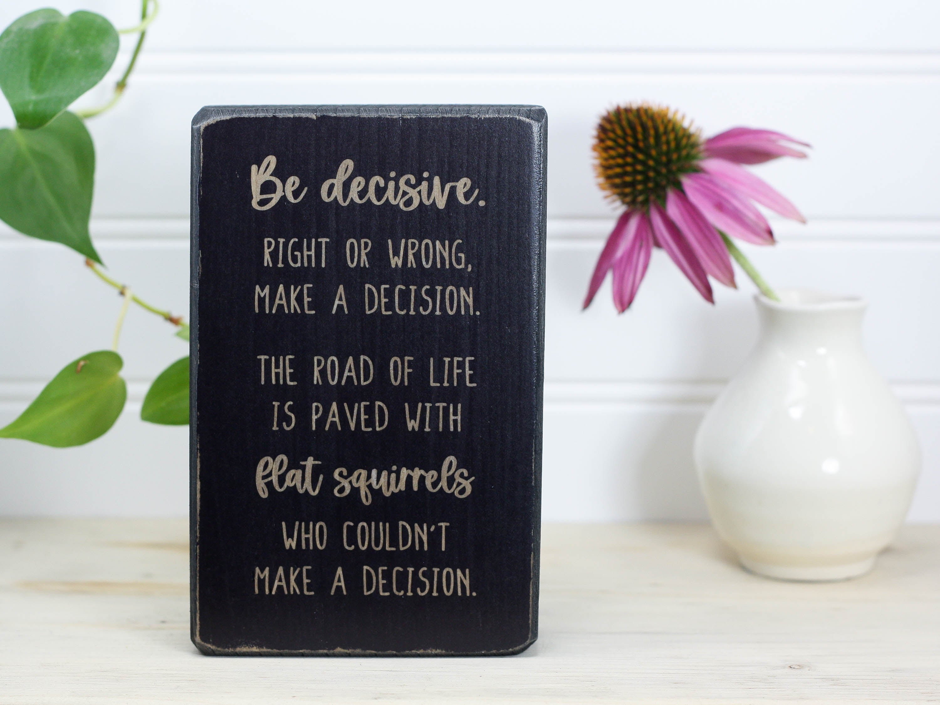 Small distressed black wood freestanding sign in with the saying "Be decisive. Right or wrong, make a decision. The road of life is paved with flat squirrels who couldn't make a decision."
