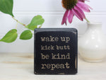 Small desk sign in distressed black with the saying "Wake up Kick butt Be kind Repeat".