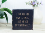 Small, freestanding, distressed black, solid wood sign with funny saying on it "I do all my own stunts, but never intentionally."