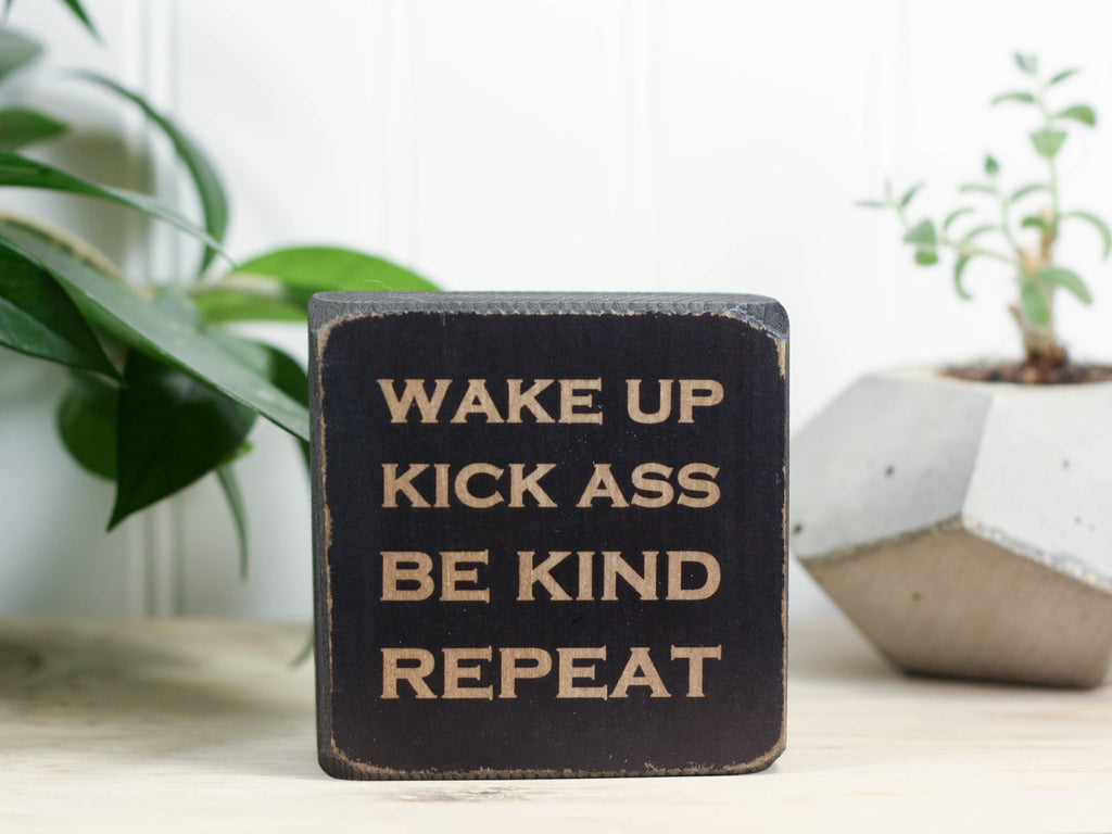 Small desk sign in distressed black with the saying "Wake up Kick ass Be kind Repeat".