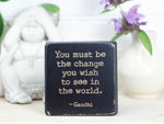 Small quote block in distressed black with the saying "You must be the change you wish to see in this world. -Gandhi"