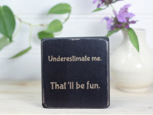 Small wood block sign in distressed black with the saying "Underestimate me. That'll be fun."