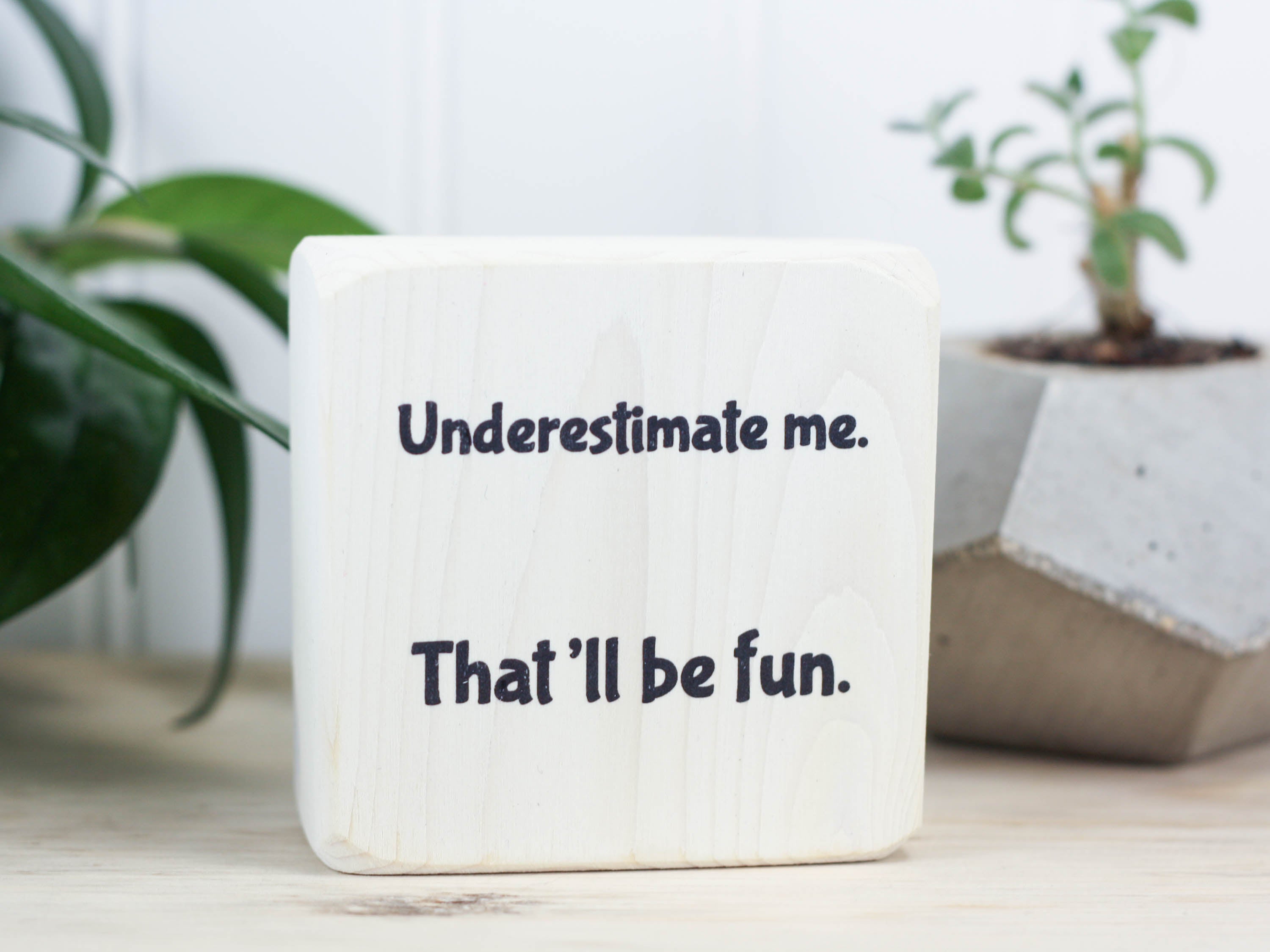 Small wood block sign in whitewash with the saying "Underestimate me. That'll be fun."