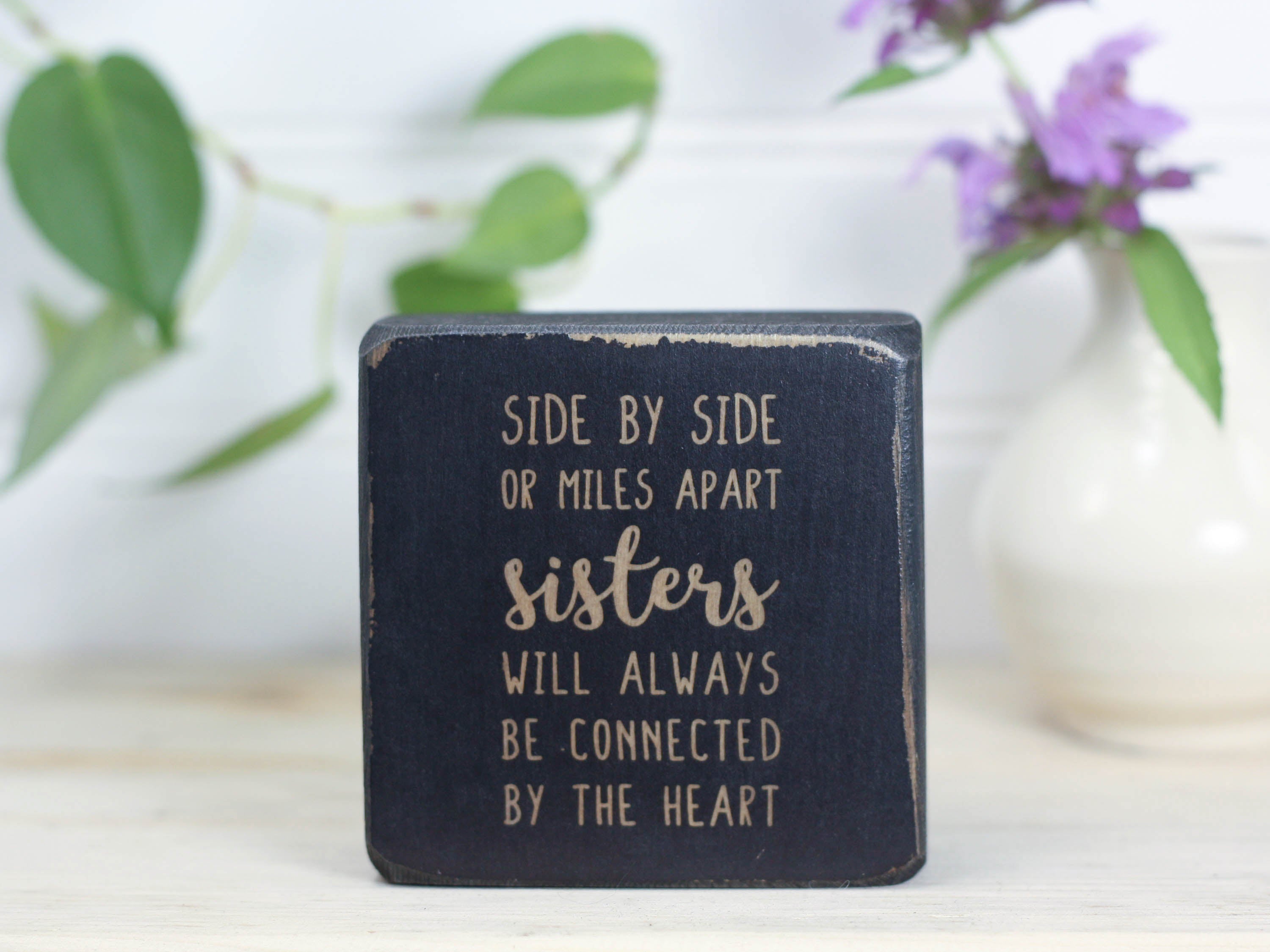 Small wood block with sister quote in distressed black that reads "Side by side or miles apart sisters will always be connected by the heart."