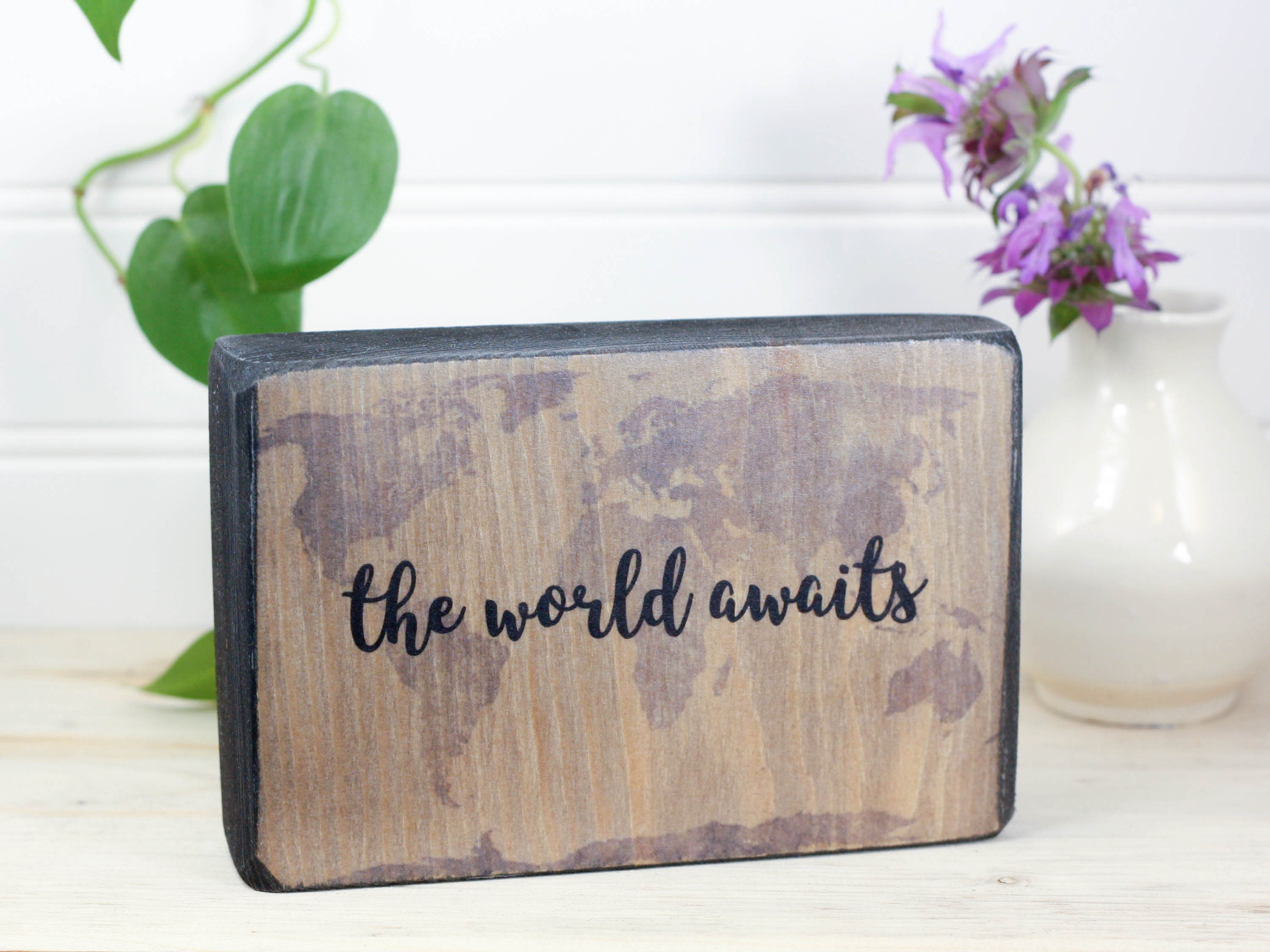 Small wood quote block in distressed black with the saying "The world awaits".