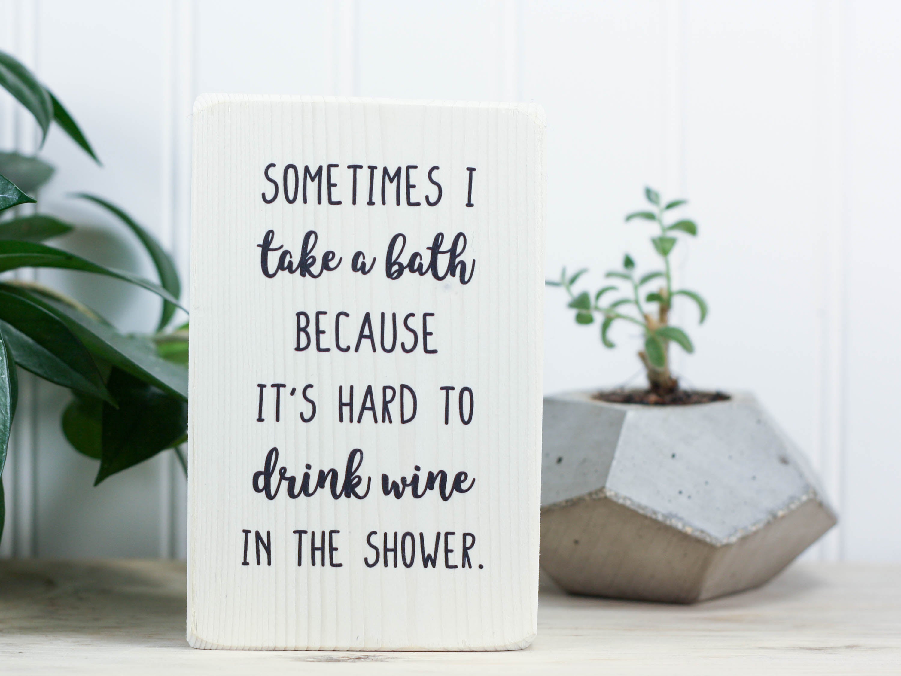 Small wood sign in whitewash with the saying "Sometimes I take a bath because it's hard to drink wine in the shower."