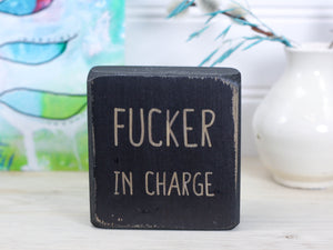 Small distressed black freestanding wood sign with the text "fucker in charge"