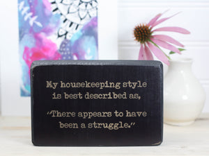Small funny wood sign in distressed black with the saying "My housekeeping style is best desribed as, "There appears to have been a struggle."
