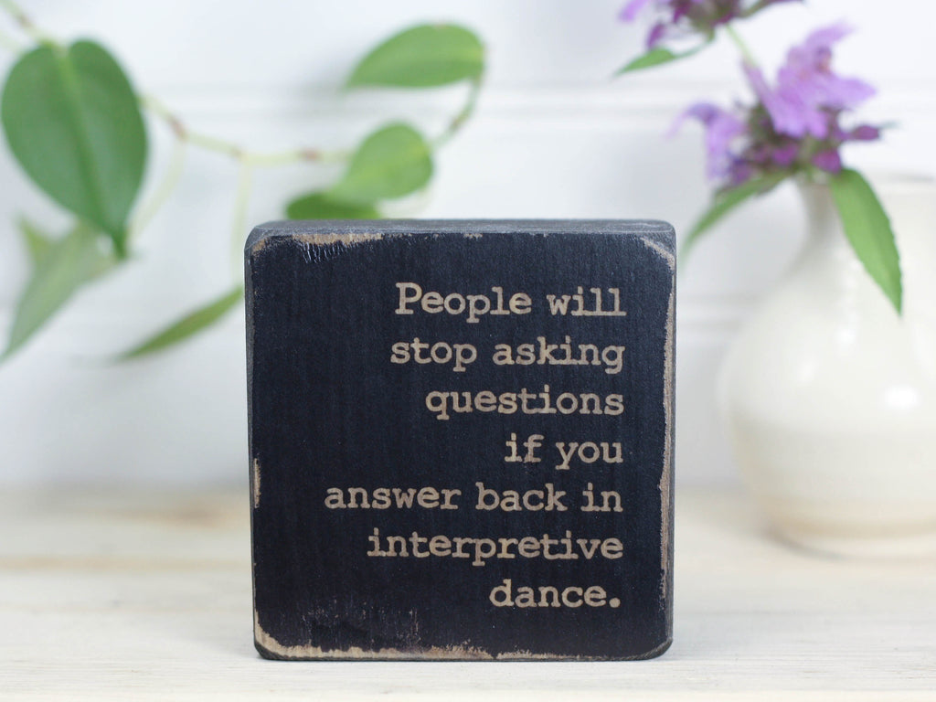 Small funny wood sign in distressed black with the saying "People will stop asking questions if you answer back in interpretive dance."